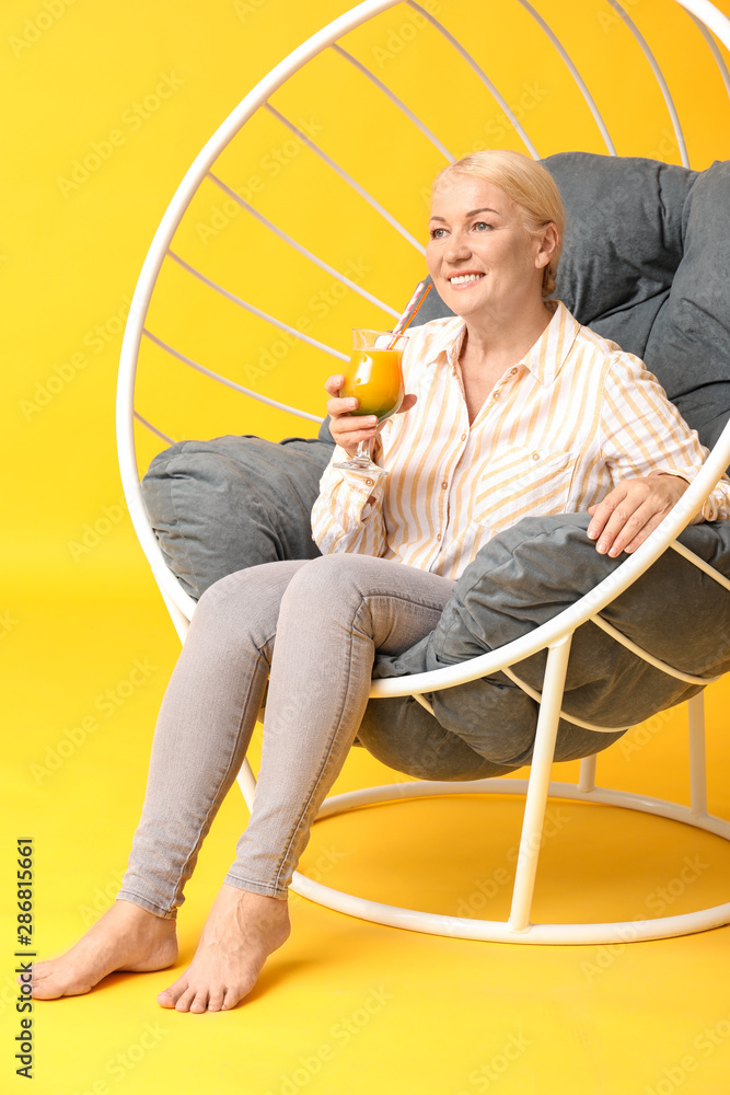 Beautiful mature woman drinking fresh cocktail in armchair on color background