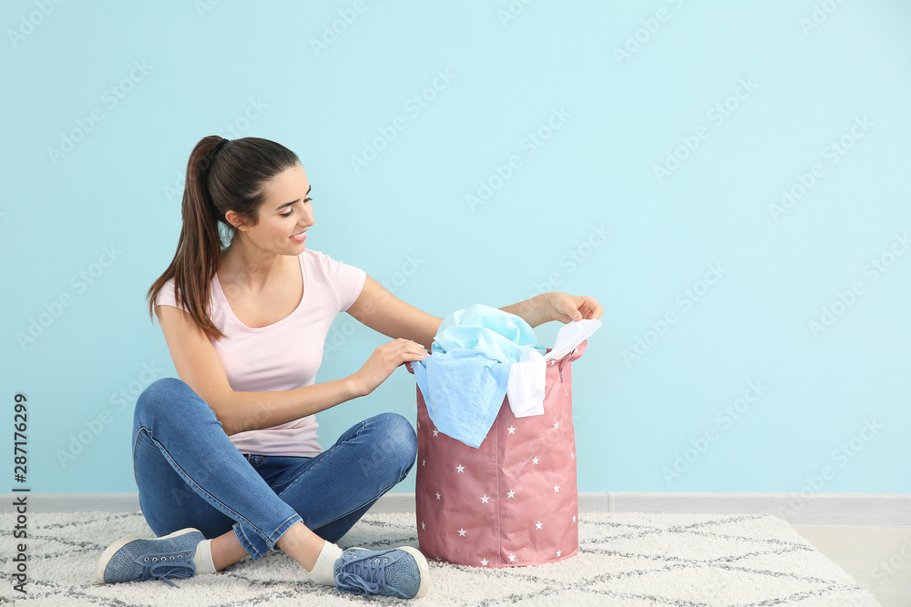 Beautiful young woman with laundry sitting on floor near color wall