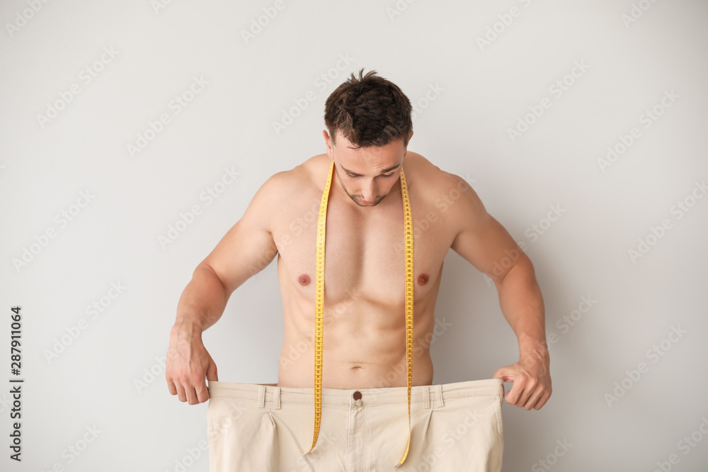 Handsome muscular man in loose pants and with measuring tape on light background. Weight loss concep