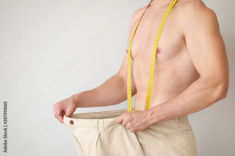 Handsome muscular man in loose pants and with measuring tape on light background. Weight loss concep