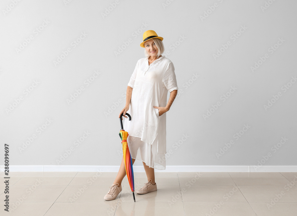 Stylish mature woman with umbrella against light wall