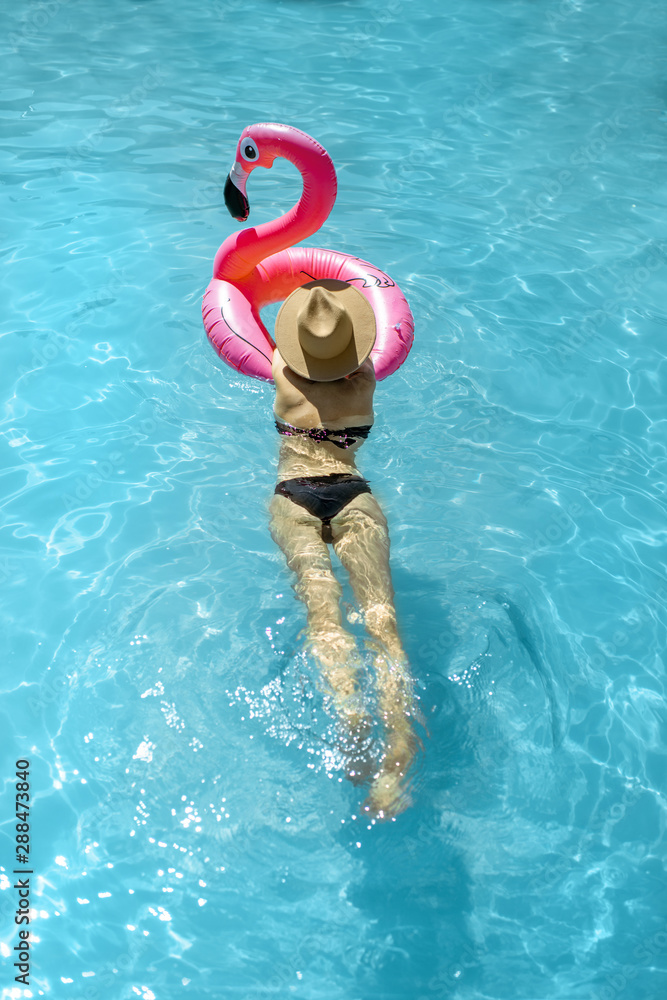 Woman in swimsuit and hat swimming with inflatable flamingo ring in the water pool, enjoying vacatio