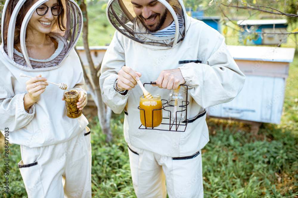 Two beekepers in protective uniform standing together with honey in the jar, tasting fresh product o