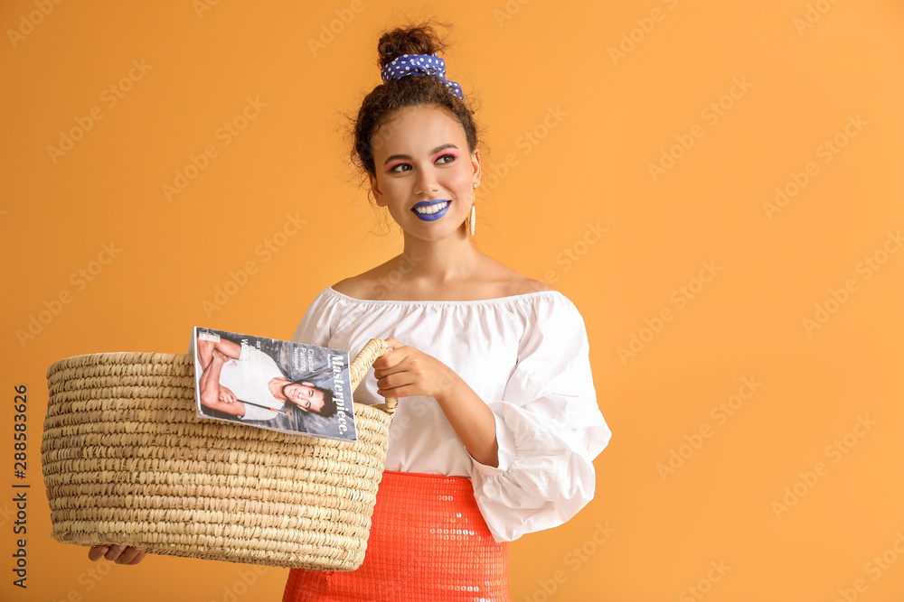 Portrait of fashionable young African-American woman with wicker basket and magazine on color backgr