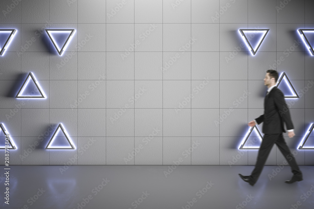 Blurred businessman walking on concrete floor along blank grey wall with triangle lights, mock up.