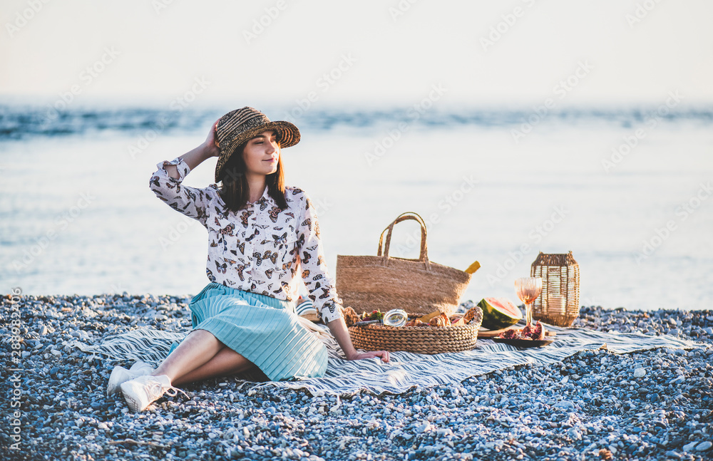 Summer beach picnic at sunset. Young woman in hat sitting on blanket and having weekend picnic outdo