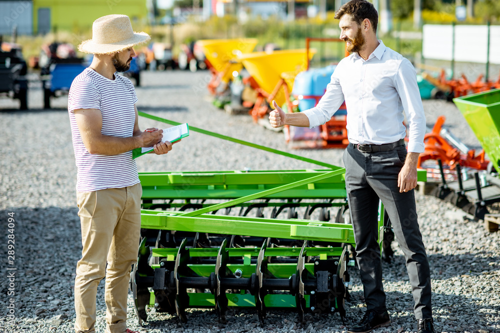 Young agronomist with salesman or manager at the open ground of the shop with agricultural machinery