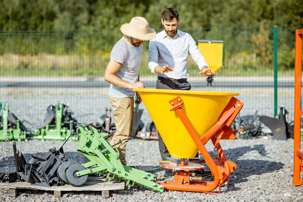 Young agronomist with salesman at the open ground of the shop with agricultural machinery, buying a 