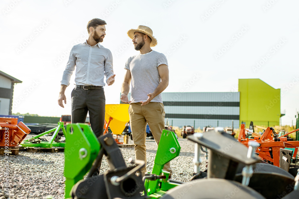 Young agronomist walking with salesman at the open ground of the shop with agricultural machinery, c