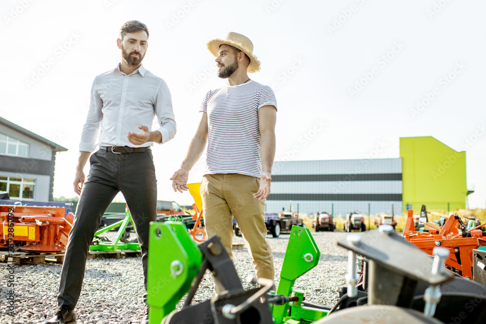 Young agronomist walking with salesman at the open ground of the shop with agricultural machinery, c