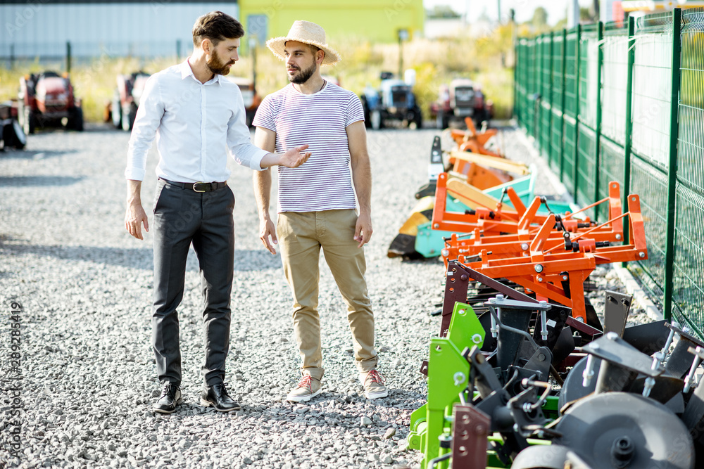 Young agronomist walking with salesman at the open ground of the shop with agricultural machinery, c