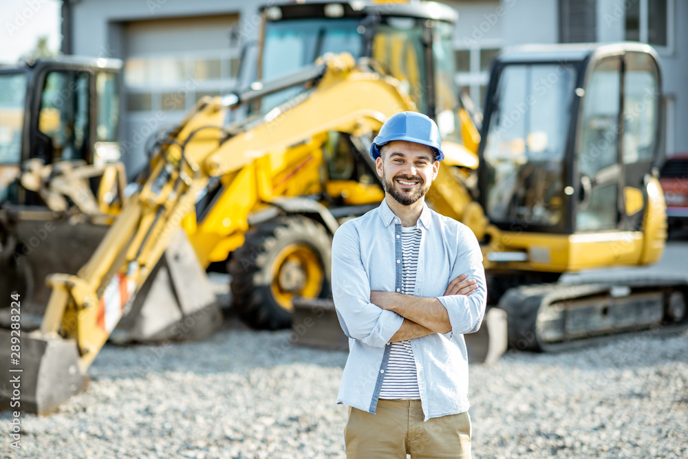 Portrait of a handsome builder standing on the open ground of the shop with heavy machinery for cons