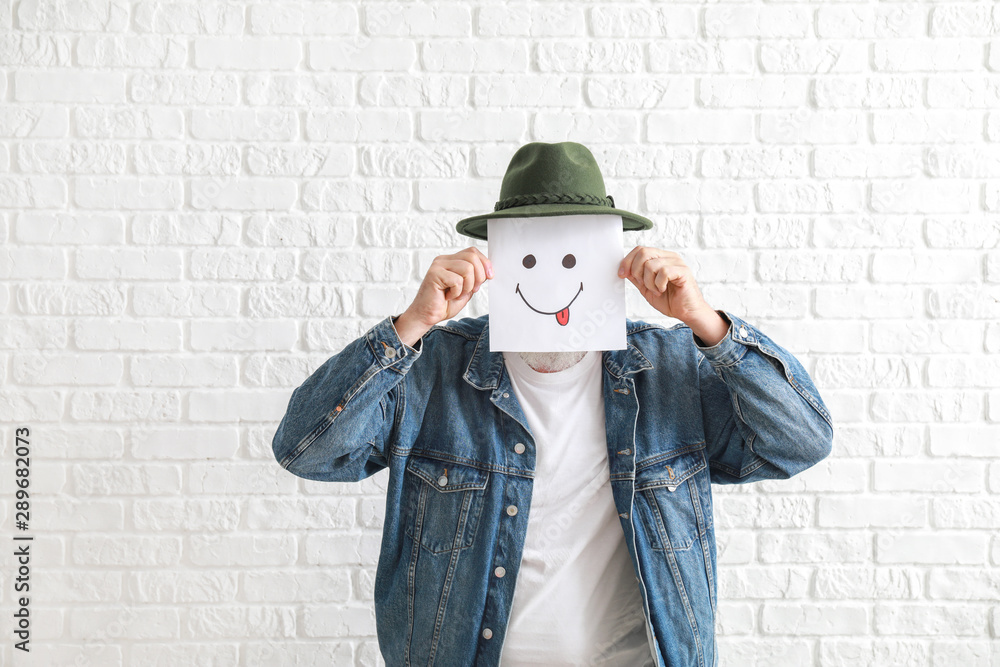 Mature man hiding face behind emoticon against white brick wall