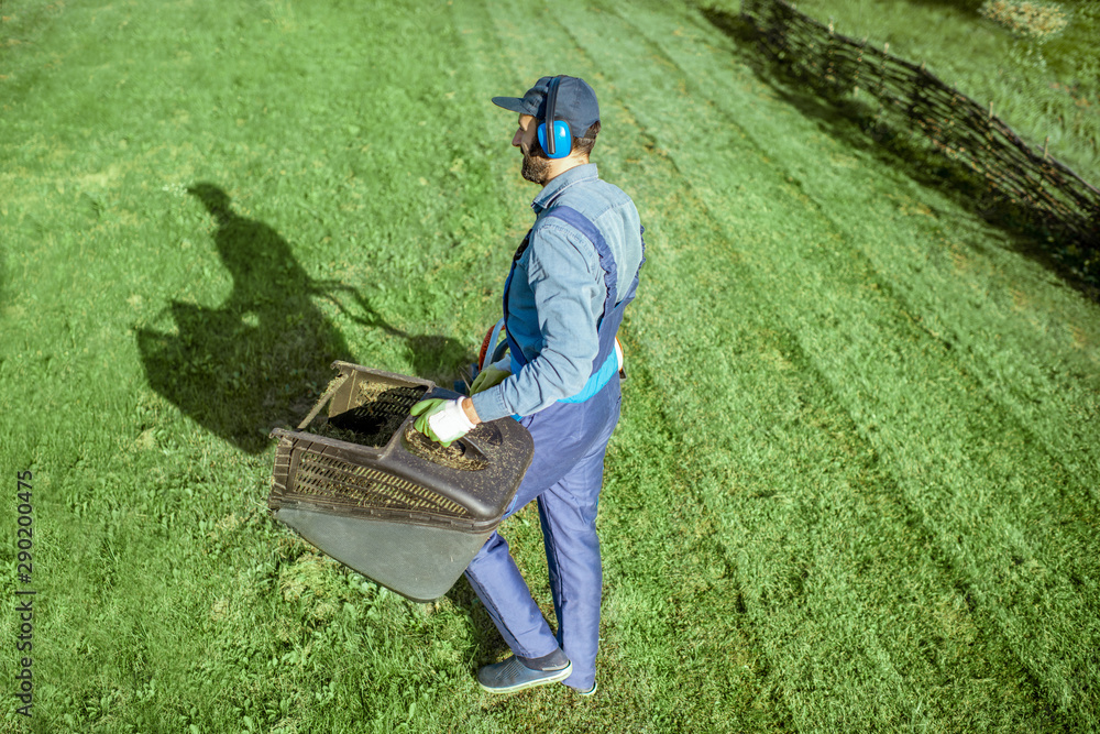 Professional gardener in protective workwear carrying basket full of grass while cutting grass with 