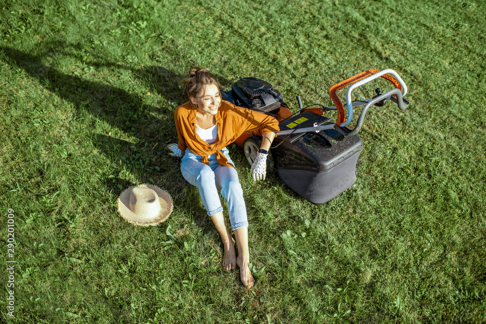 Beautiful young woman resting on the grass near the lawn mower enjoying gardening on the backyard in