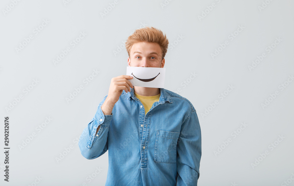 Young man holding paper sheet with drawn smile on light background