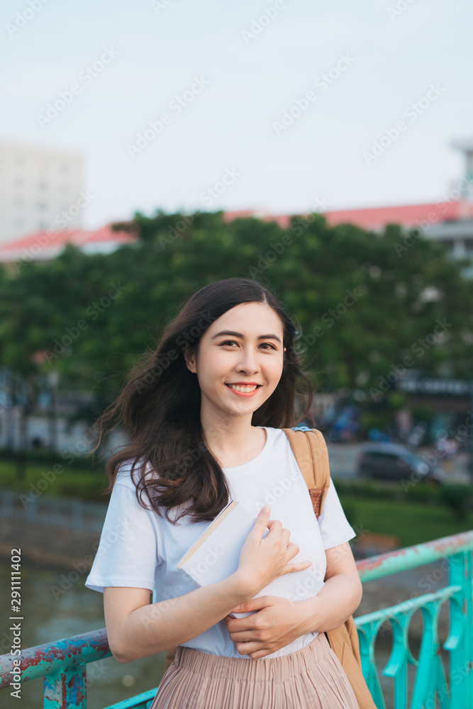 美丽的亚洲女大学生拿着书站在户外，进行人文教育学习