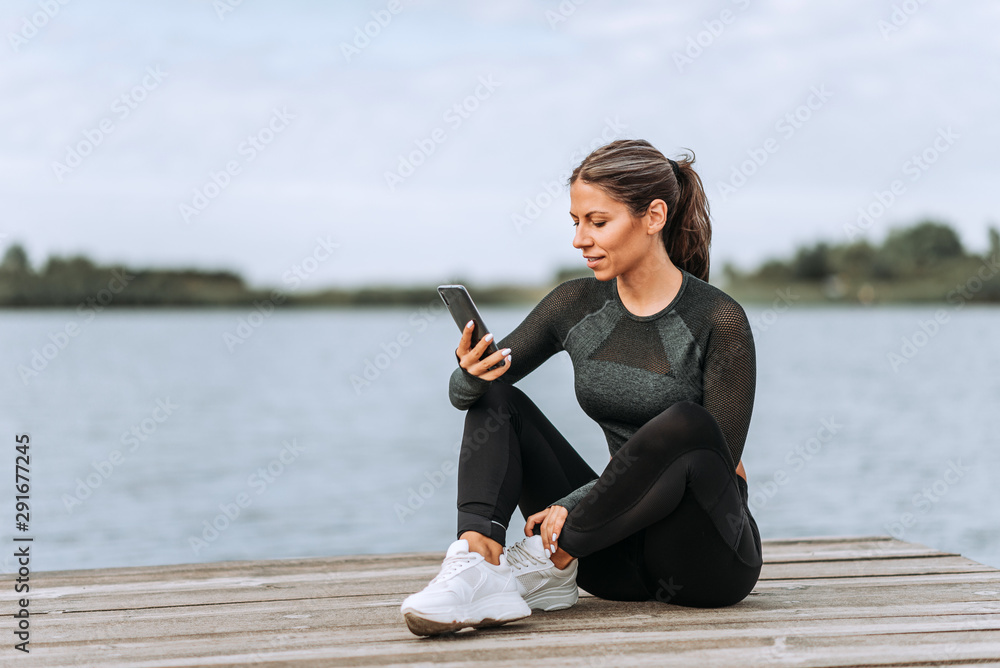 在河边放松的运动女性，正在使用手机。