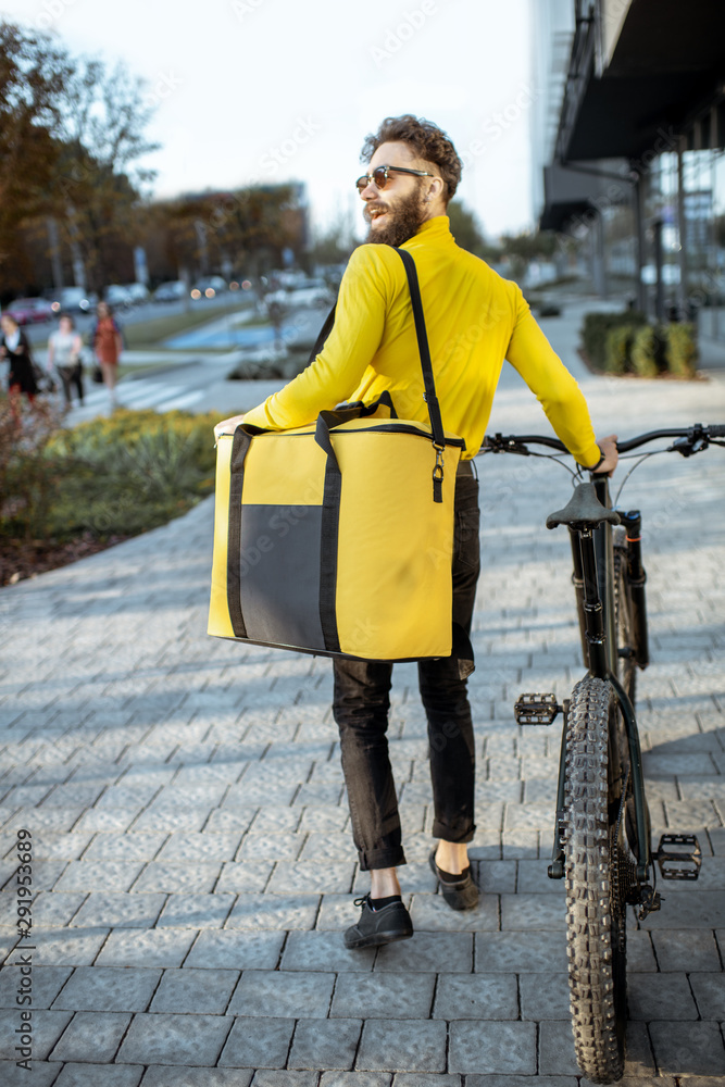 Young courier delivering food in a yellow thermo bag, walking with bicycle on the street in the city