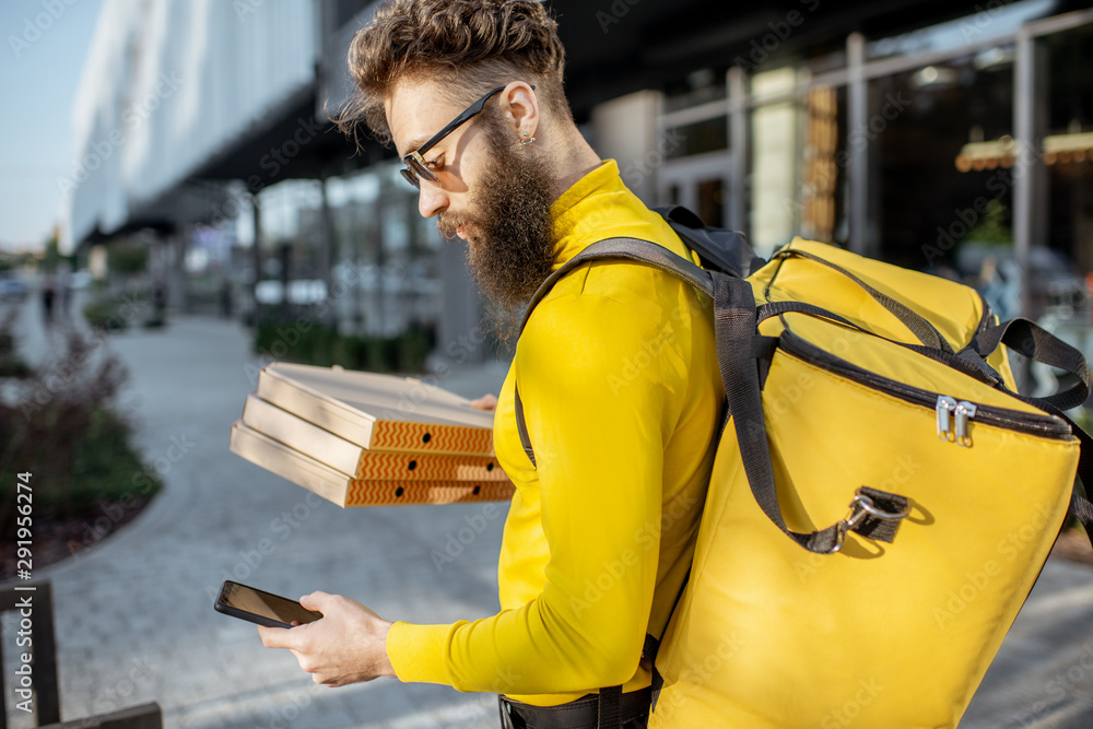 Young male courier in yellow sweatshirt delivering pizza, standing with thermal backpack and smart p