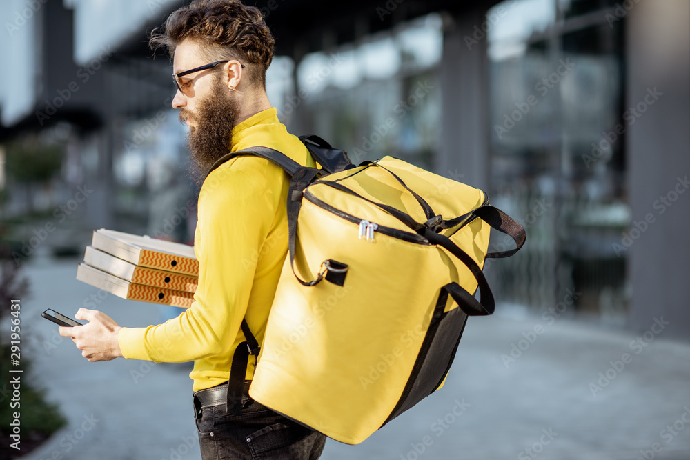 Young male courier in yellow sweatshirt delivering pizza, standing with thermal backpack and smart p