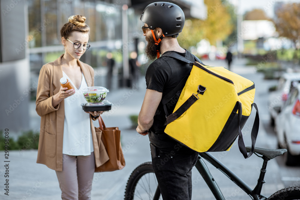 Courier delivering fresh lunches to a young business woman on a bicycle with thermal backpack. Takea