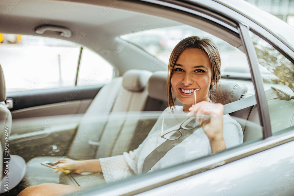 Portrait of a positive young female entrepreneur sitting on a back seat of a car and smiling at came