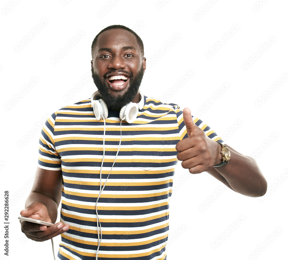 Happy African-American man with mobile phone and headphones showing thumb-up gesture on white backgr