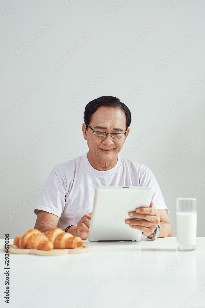 Elder man drinking milk and reading news on his digital tablet instead of newspaper