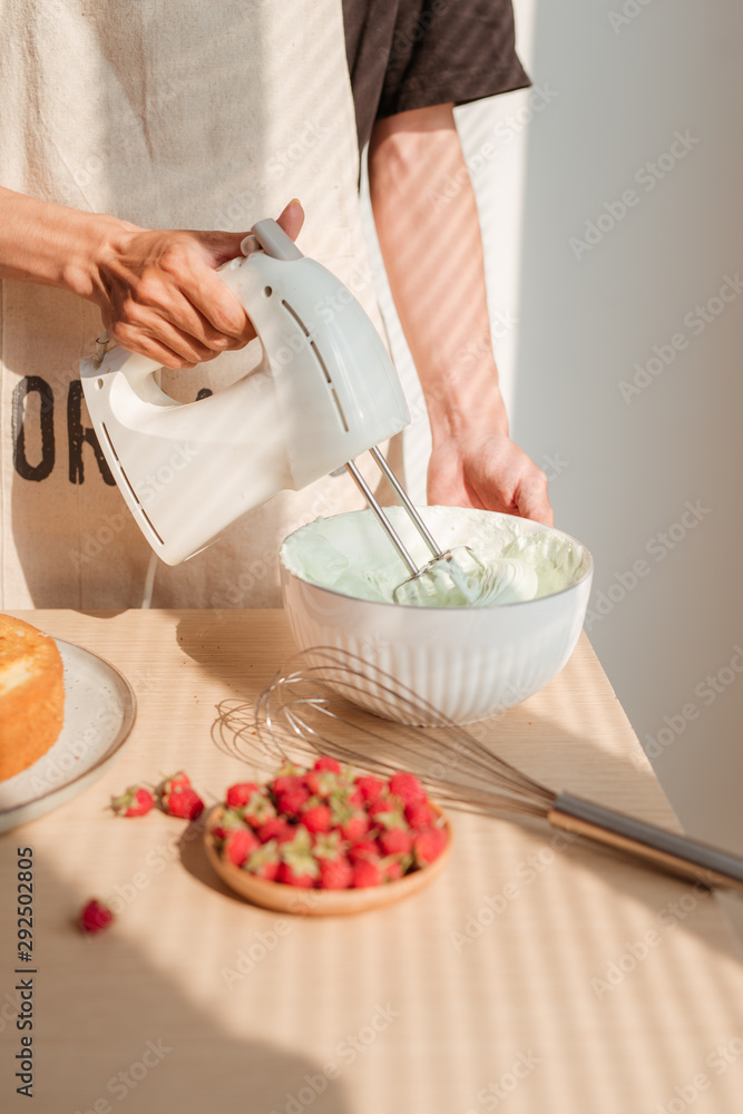 Male hands whipping whites cream in glass bowl with mixer on wooden table. Making sponge cake or red