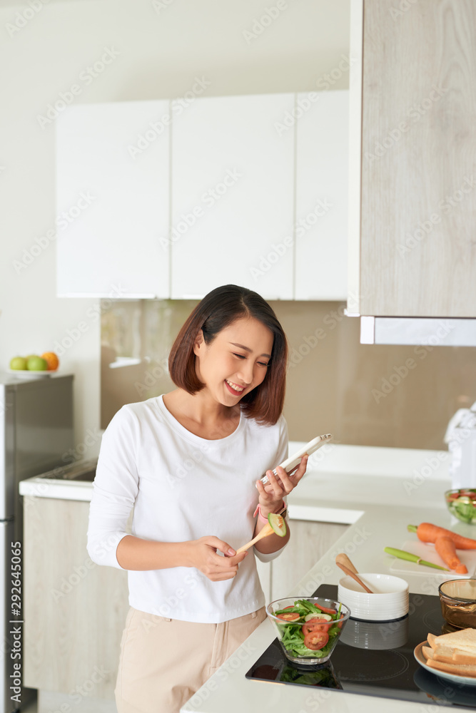 漂亮的年轻女人穿着围裙在家里的厨房里用手机烹饪健康沙拉