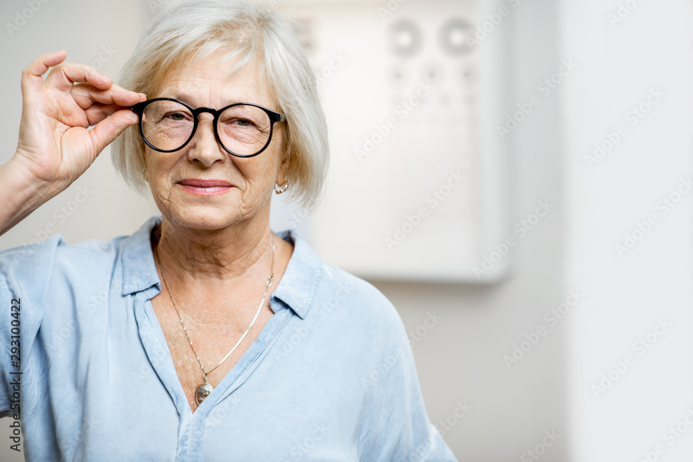 Portrait of a happy senior woman wearing eyeglasses in front of eye chart in ophthalmology office. C