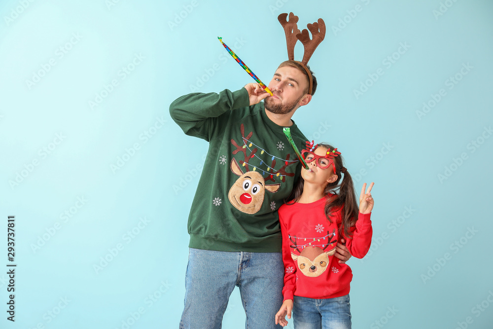 Funny little girl and her father in Christmas sweaters and with party whistles on color background