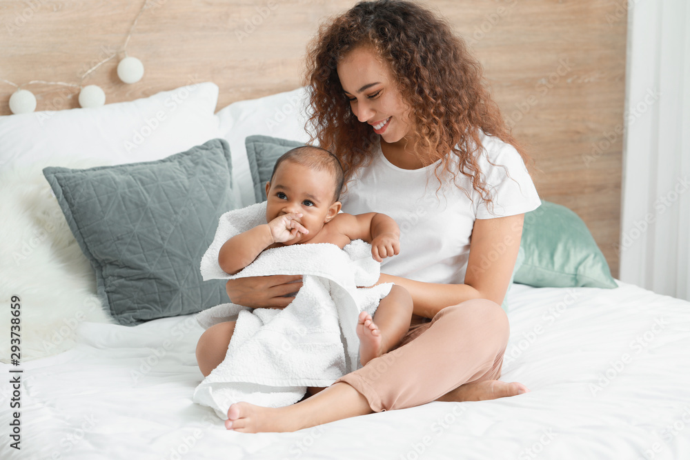 Young African-American mother with cute baby sitting on bed after bathing