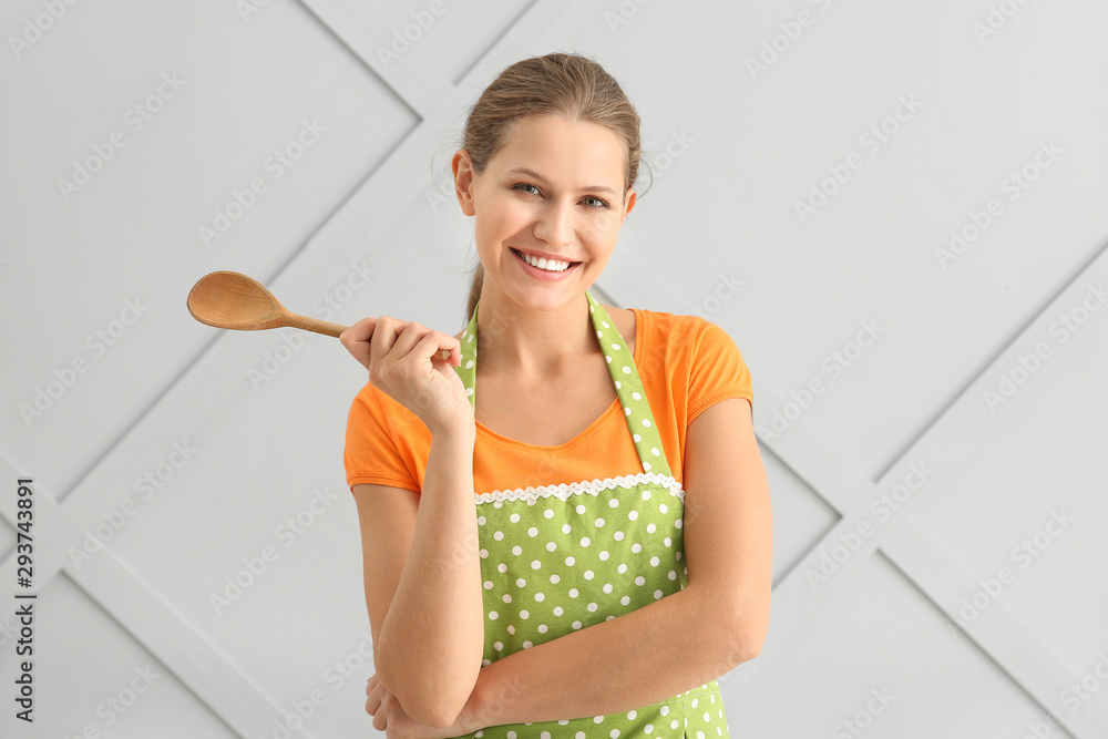 Beautiful young woman in apron and with spoon on light background