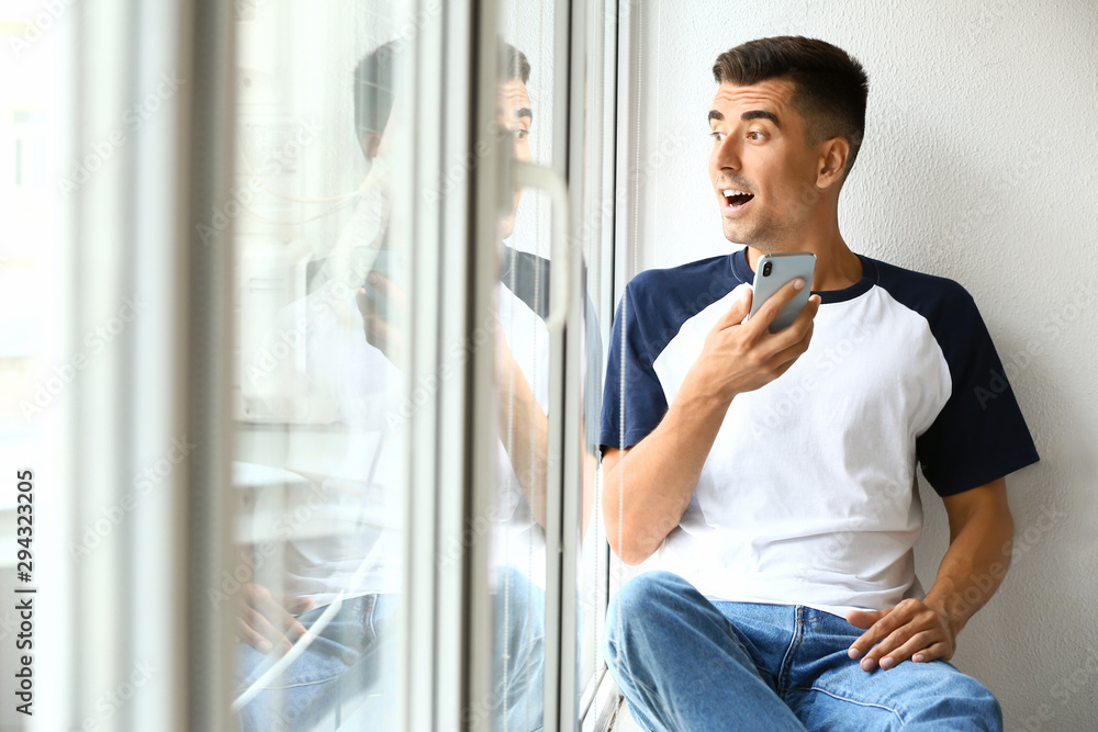 Surprised young man with mobile phone near window