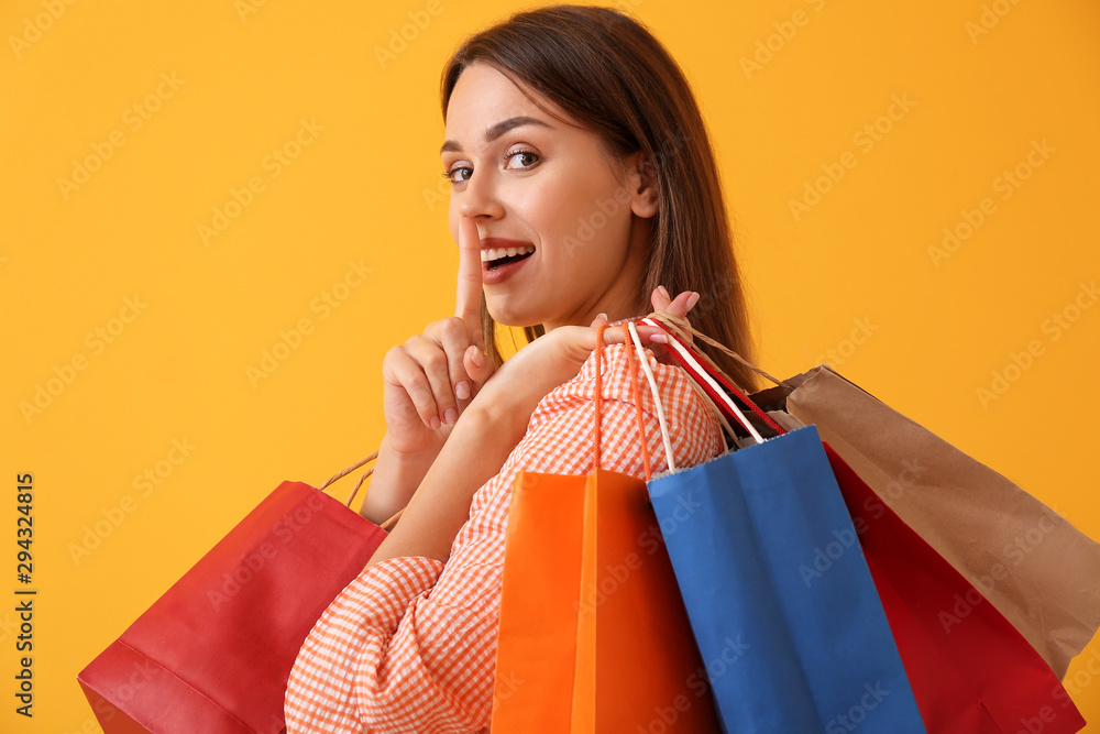 Beautiful young woman with shopping bags showing silence gesture on color background