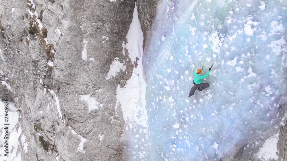 AERIAL: Female climber placing her ice-picks while climbing up an icy waterfall.