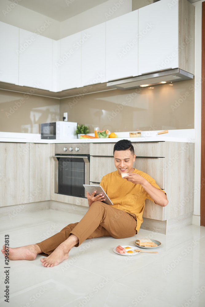 Asian young man work at home, an owner of online business, eating .sausage and looking on tablet.