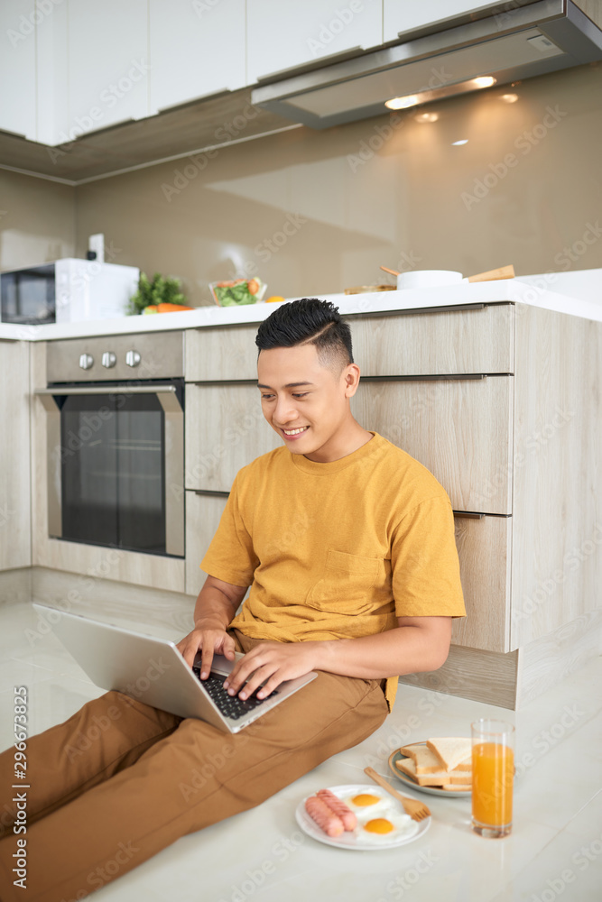 attentive asian man using laptop while sitting on floor in kitchen