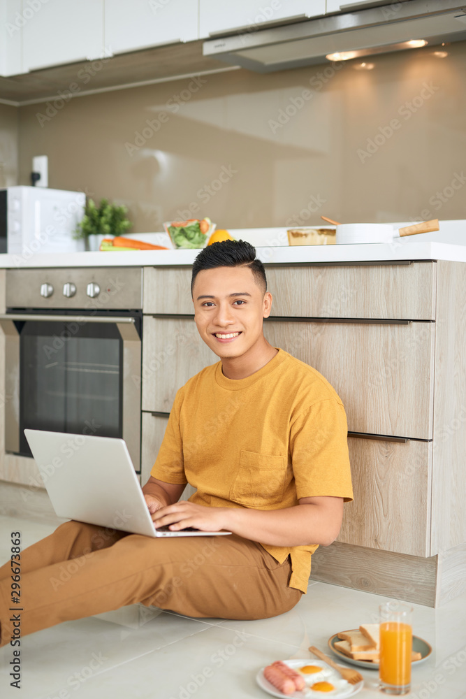 Young business man is smiling, eating breakfast and working on laptop in bright kitchen.