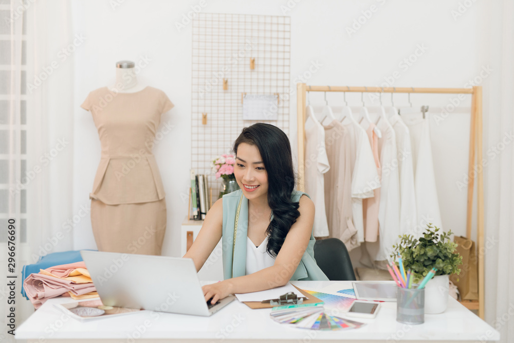 Portrait of young Asian fashion designer sitting at the desk in front of her laptop in the workshop