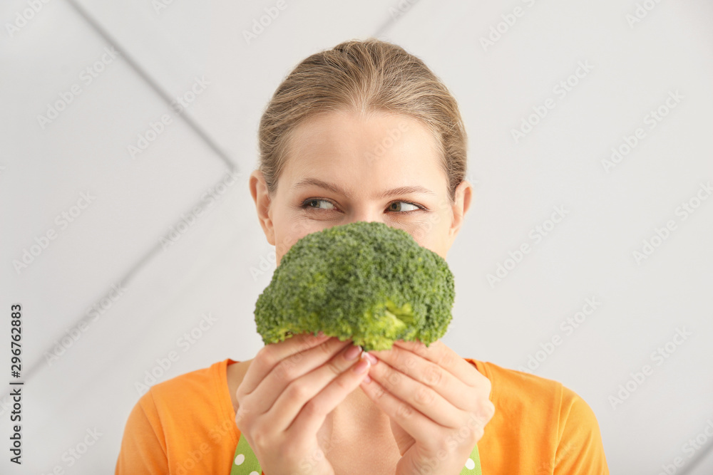 Beautiful young woman in apron and with broccoli on light background