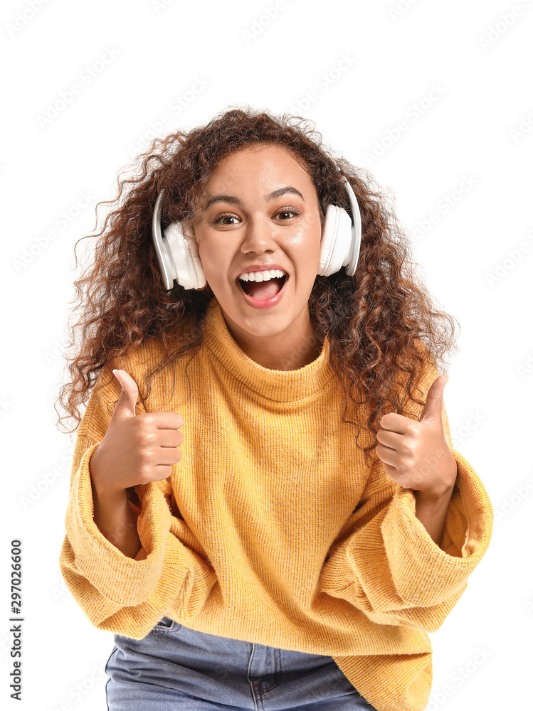 Beautiful African-American woman showing thumb-up while listening to music on white background