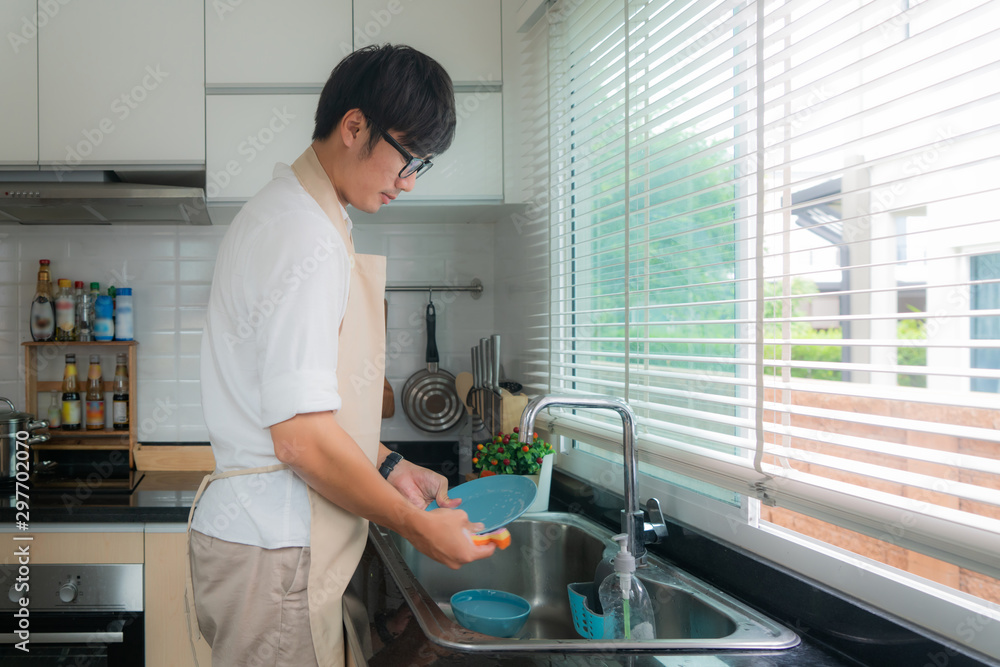 Happy Asian young man in white shirt standing and washing dishes in sink on the kitchen. Kitchen orn