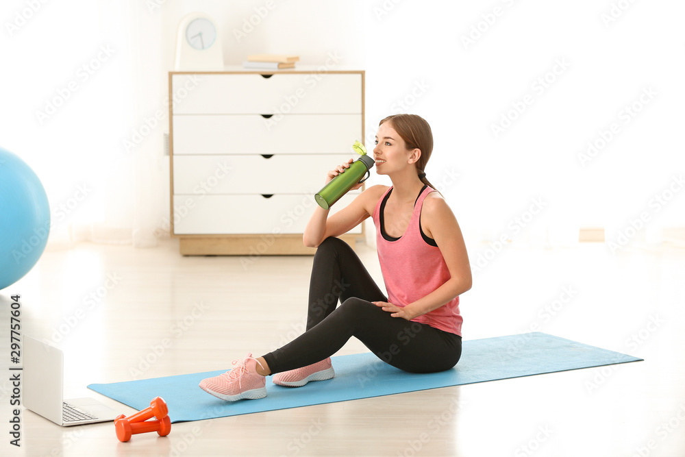 Sporty young woman with laptop and bottle of water at home