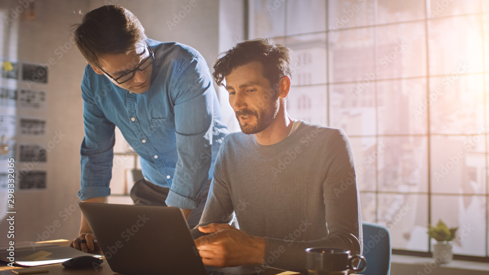 Professional Creative Man Sitting at His Desk works on a Laptop, His Colleague Helps with Poignant A