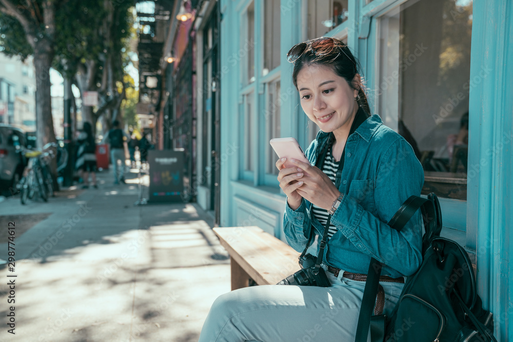 时尚的亚洲女孩背包客坐在sto外城市步道的长椅上使用手机应用程序