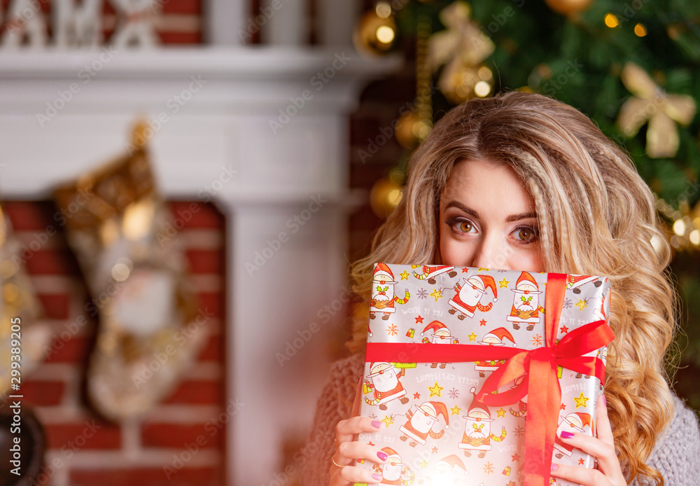 Girl in a sweater near the Christmas tree with a gift near face, New Year Christmas tree. Young woma