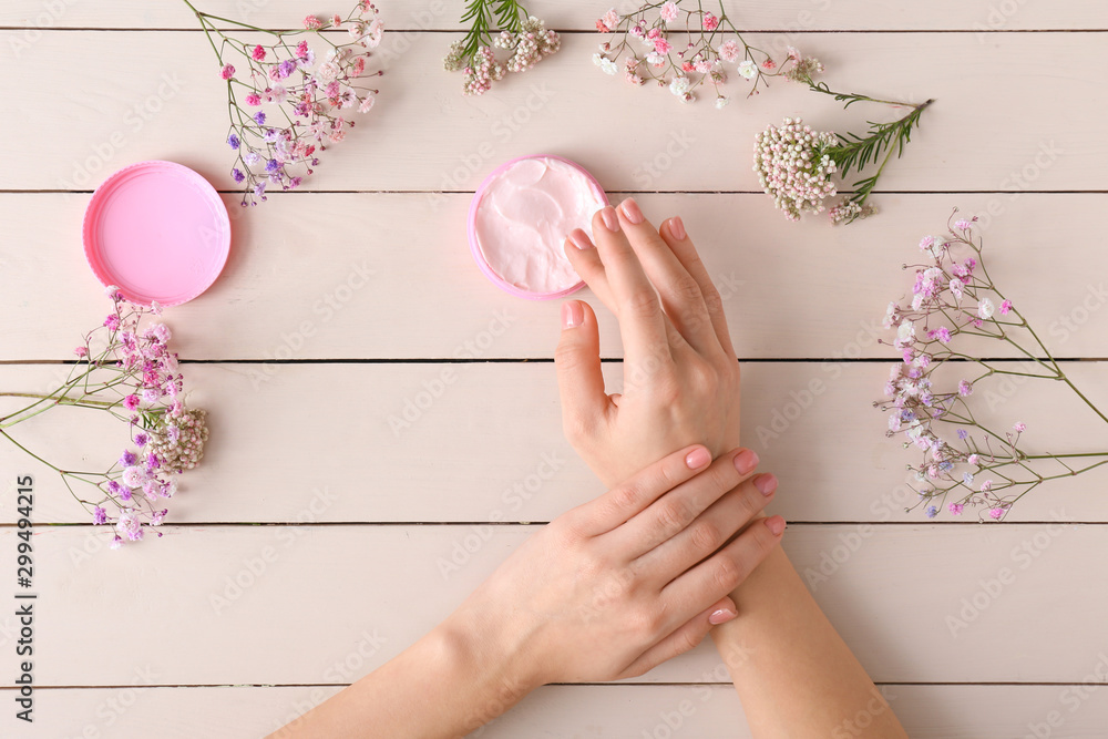 Hands of young woman, jar of cream and flowers on wooden background